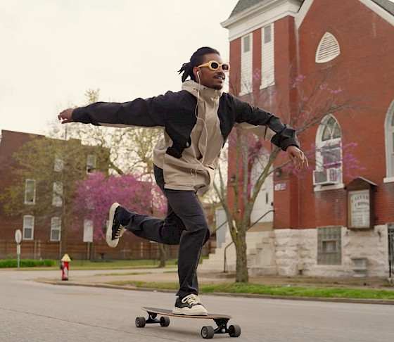 Video still of a man wearing a black and white hoodie, black pants, black sneakers, and white sunglasses rolling by on a skateboard on an asphalt road in front of a red brick church.