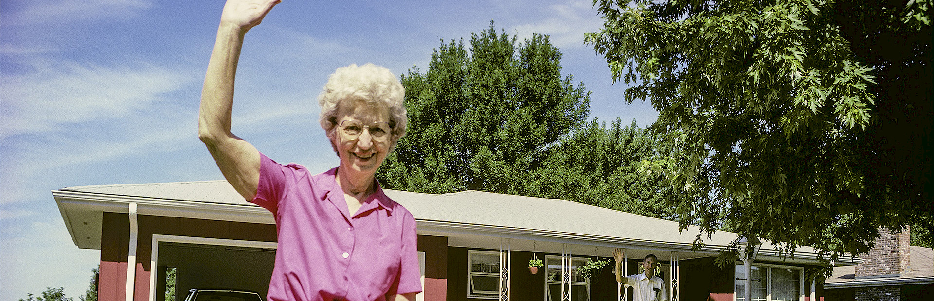Photo of a woman standing in the driveway of a red house waving.