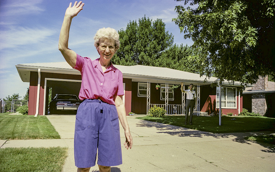 Photo of a woman standing in the driveway of a red house waving.
