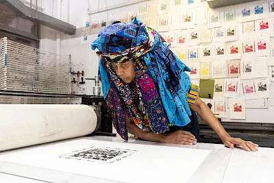 Photo of Chakaia Booker wearing a large blue headress in a workshop looking down at a print of her work on table in the foreground. There are dozens of color samples of a print on the wall behind her.