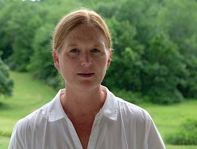 Headshot of Anne Lindberg wearing a white collared shirt with her red hair pulled back standing in front of a green background of grass and trees.