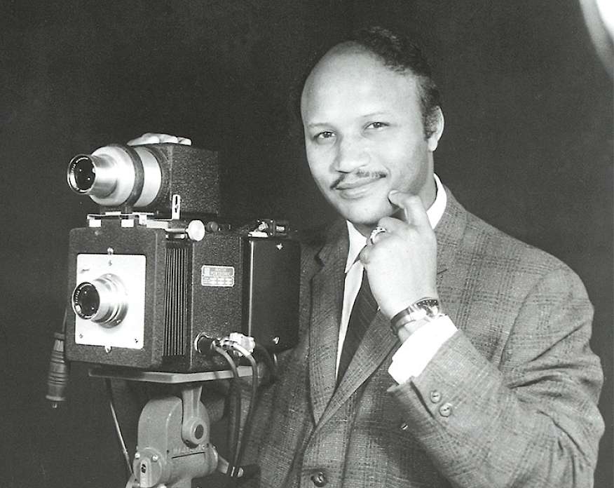 Black and white self-portrait photograph of Michael Grice wearing a suit and standing next to an antique camera.