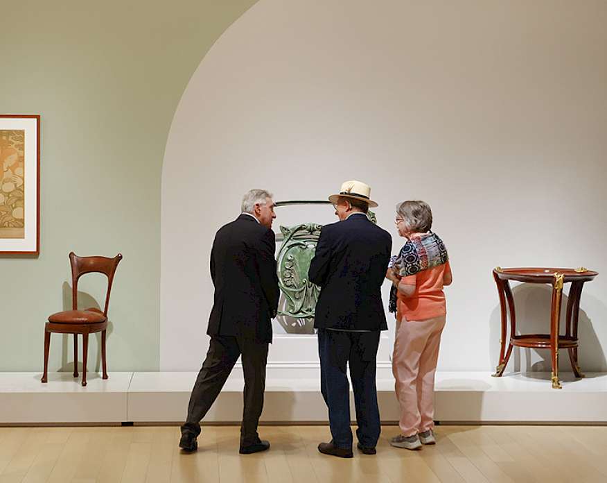 Photo of two men and a woman looking at Art Nouveau furniture in an art gallery.