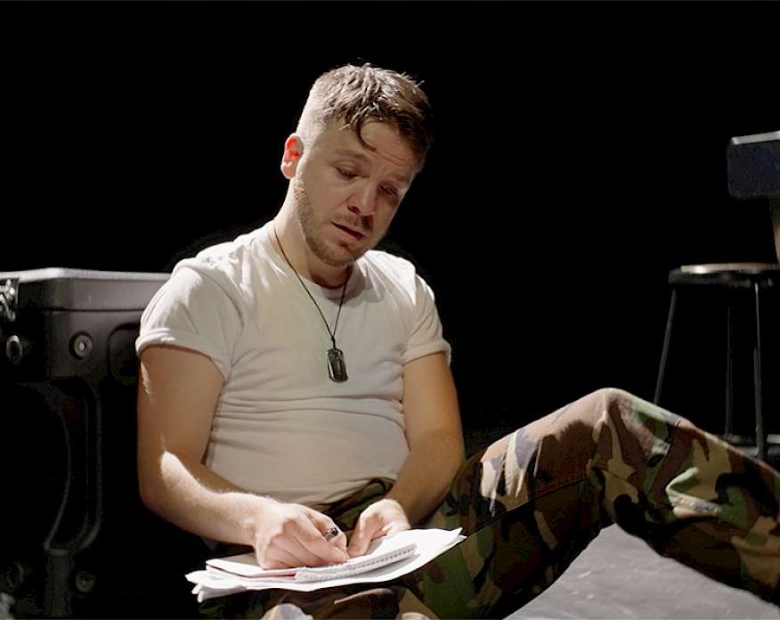 Photo of a man wearing a black t-shirt and military beret looking at his feet on a stage in front of a project screen displaying a close-up photo of a military bracelet on an arm.