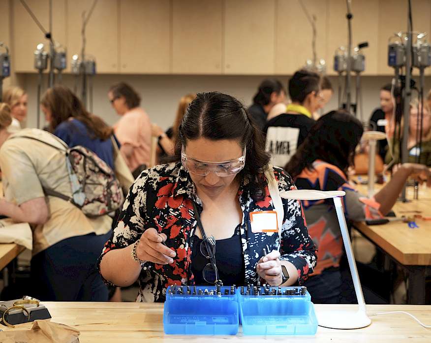 Photo of a woman wearing goggles and looking in a blue box of metal tools in an art studio where other people are interacting in the background.