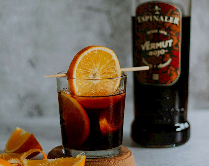 Photo of a dark brown cocktail garnished with orange in a rocks glass sitting next to a bottle of vermouth.