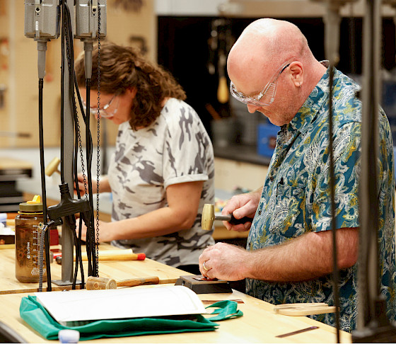 Photo of two people wearing goggles and hammering metal in an art studio.