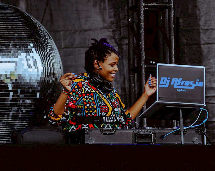 Photo of a female DJ wearing a brightly patterned shirt and standing next to a large disco ball behind a black DJ booth. The backside of the laptop in front of her reads DJ Afrosia.