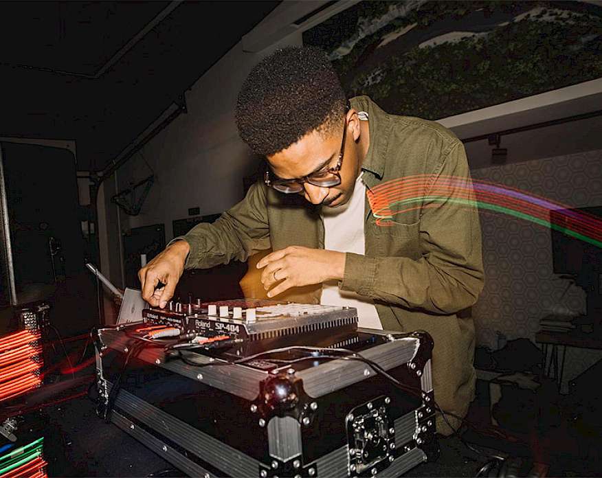 Photo of a male DJ wearing a green jacket and glasses mixing music on a soundboard set on top of a music case.