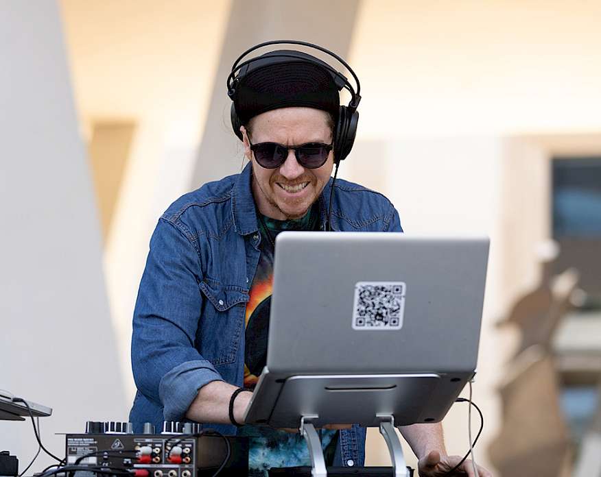 Photo of DJ Mike Poe wearing headphones and sunglasses while typing on a laptop behind a DJ booth.