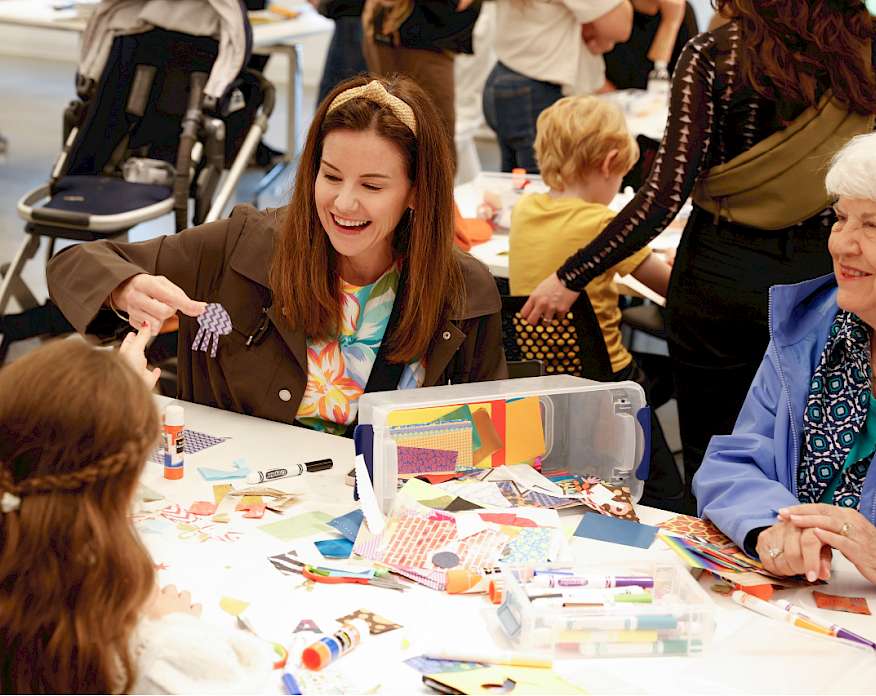 Photo of a mother, grandmother, and young girl sitting at a table doing arts and crafts with other families in the background.