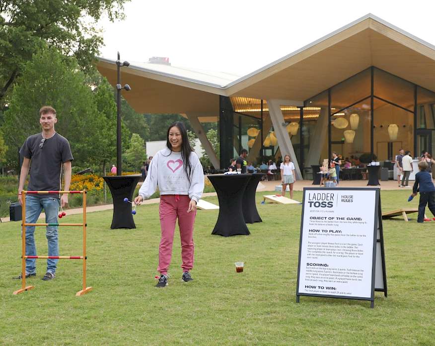Photo of a man and woman playing ladder toss on a large outdoor lawn space with other people playing other lawn games in the background.