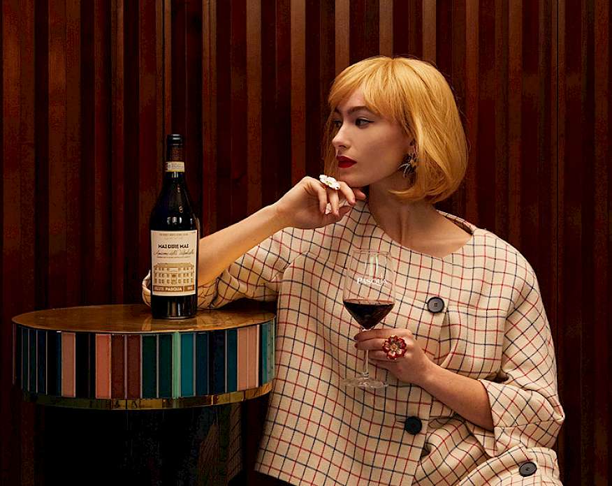 Photo of a woman holding a glass of red wine with her right elbow leaning on a golden end table where an open bottle of wine is sitting.