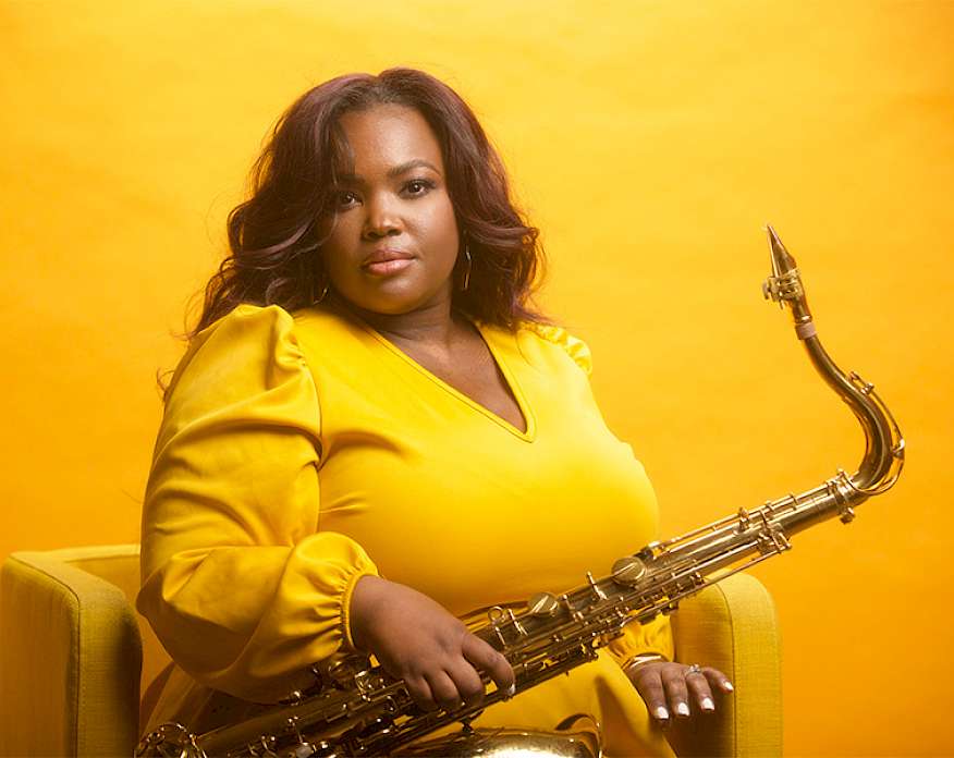 Photo of Camille Thurman wearing a yellow dress and holding a saxophone while seated in a yellow chair in front of a yellow wall.