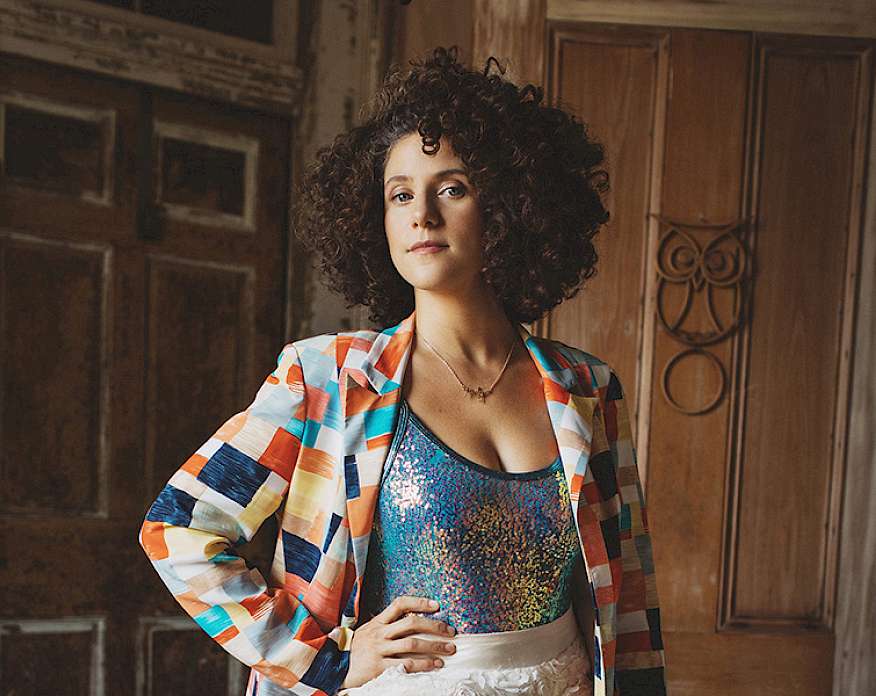 Photo of a woman with brown curly hair wearing a multi-colored jacket, sequin top, and white shorts posed with her hand on her hip.