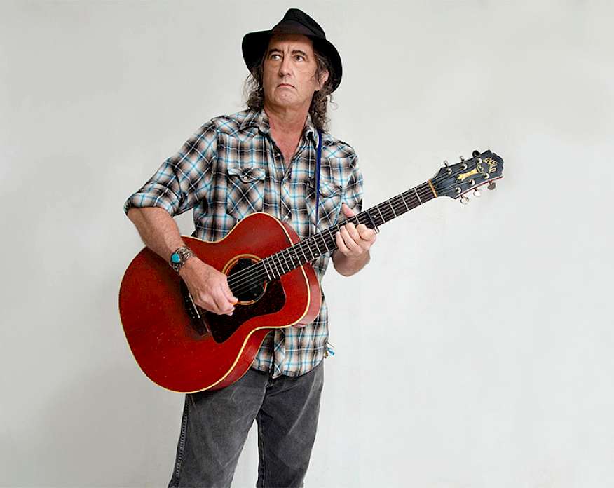 Photo of James McMurty wearing a plaid shirt and black fedora while holding an acoustic guitar in front of a light gray backdrop.