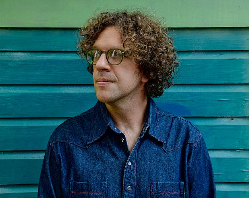Photo of Phil Cook wearing a denim shirt and glasses standing in front of a wall with blue siding.