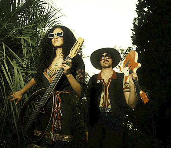 Photo of a man and woman wearing sunglasses and holding guitars while walking next to tropical trees.