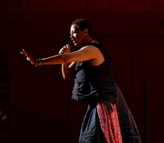 Photo of Lisa Fischer on stage under a spotlight and holding a microphone with her left arm outstretched toward the audience.