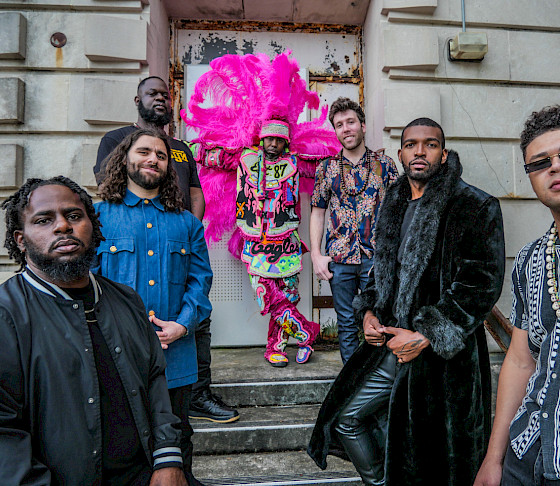 Band photo of seven men standing outside a stone building. One of them is wearing a bright pink headdress.