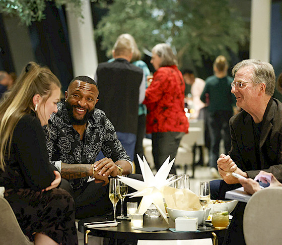 Photo of two men and two women sitting an a table drinking champagne with other people in the background.