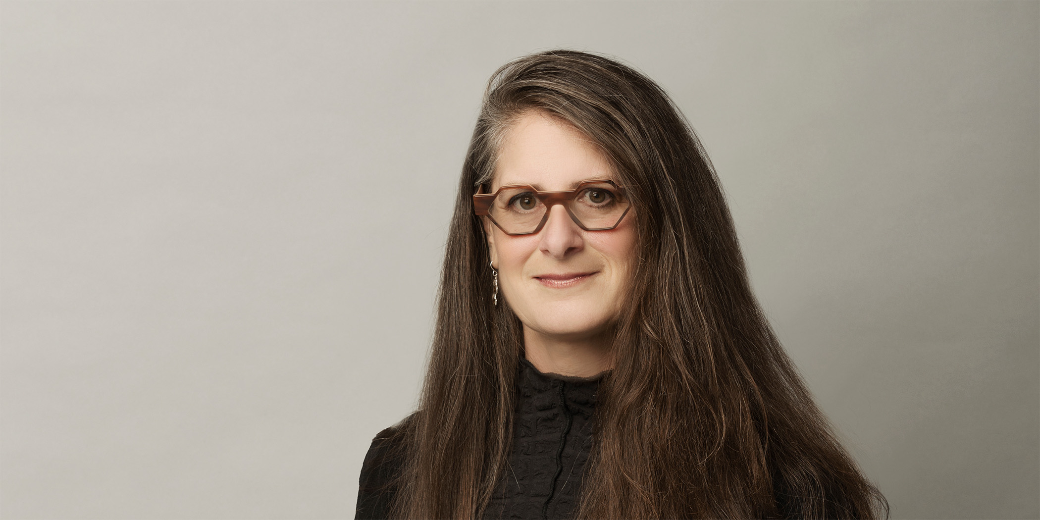 Photo of a woman with graying long brown hair wearing a black dress and thick brown frame glasses.