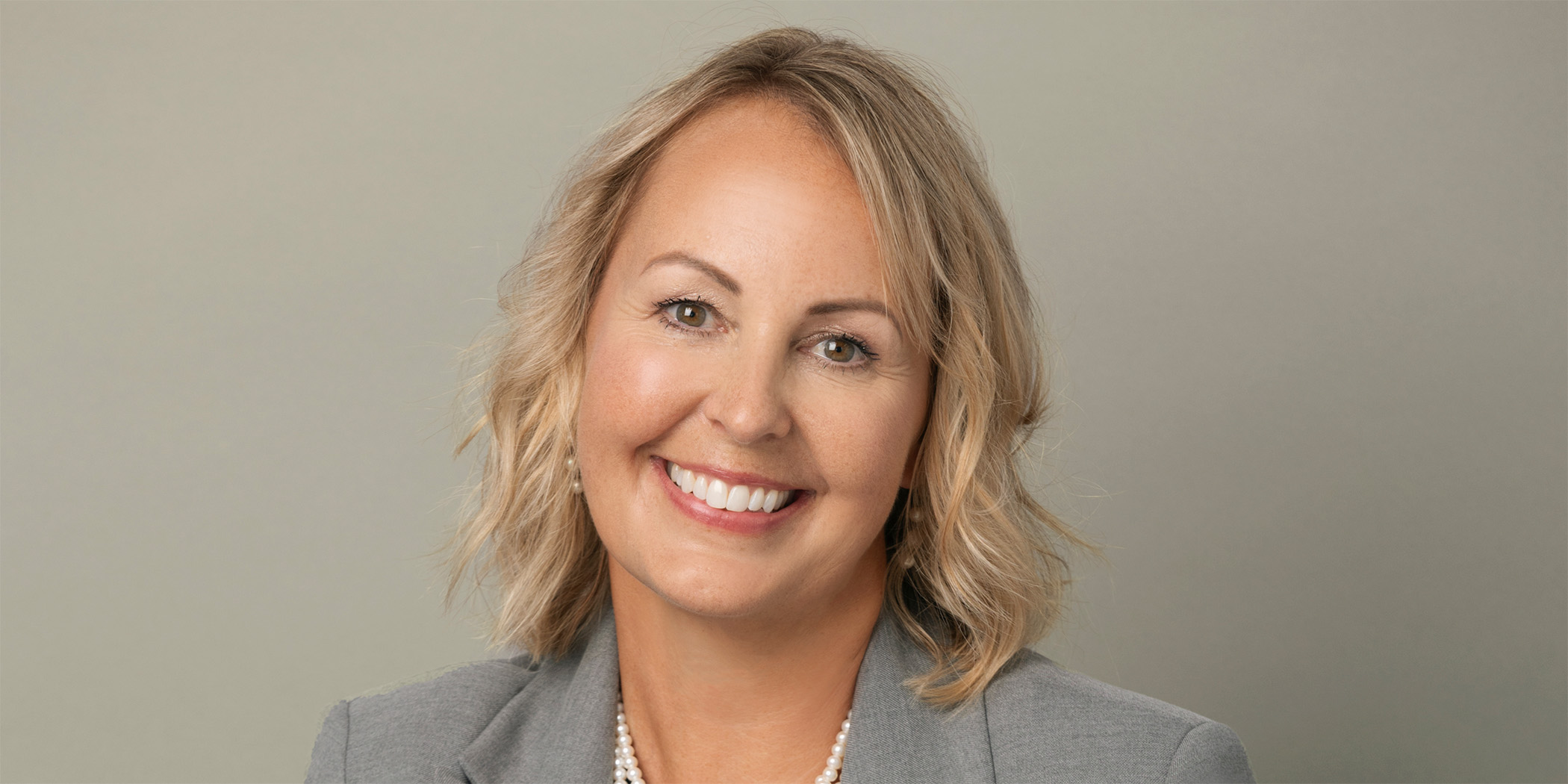 Photo of a smiling woman with short blonde hair wearing a gray blazer and a pearl necklace.