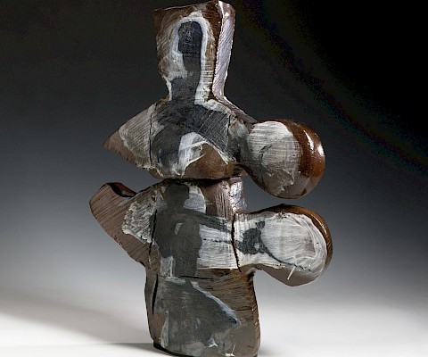 Photo of a brown, gray, and white ceramic sculpture in the shape of an abstract cross standing in front of a black background.