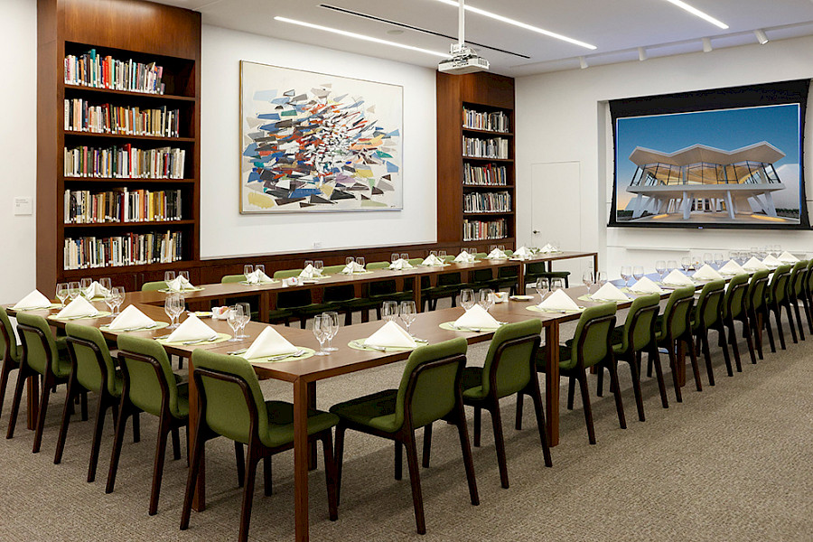 Photo of a large U-shaped arrangement of wooden tables and green fabric chairs arranged in front of floor-to-ceiling bookshelves and a projector screen.