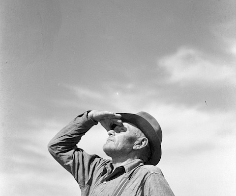 Black and white photograph of a farmer wearing a cowboy hat looking into the distance with his hand shielding his eyes from the sun.