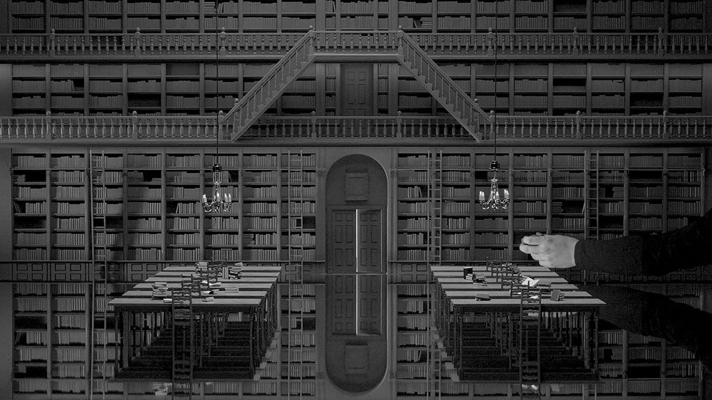 Film still depicting a hand inserting a small book into a small bookshelf on a video set decorated as a library.