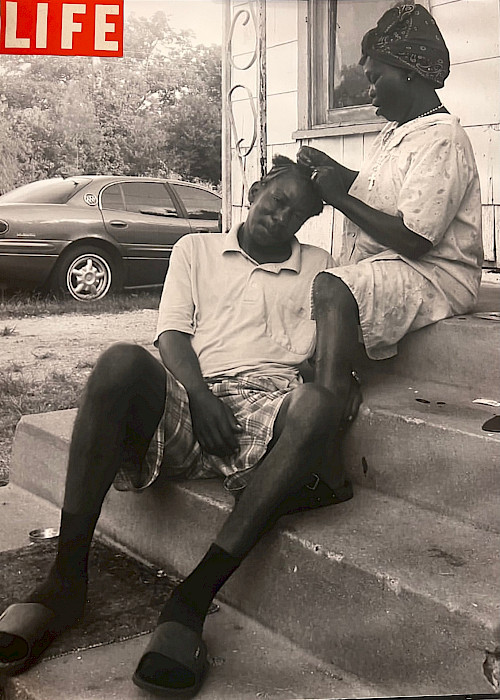 Black and white photo of a Black man seated on a porch while a seated Black woman behind him braids his hair.