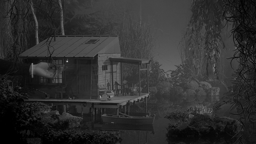 Film still depicting a hand hanging vines from a small set decorated with a boathouse next to a swamp.