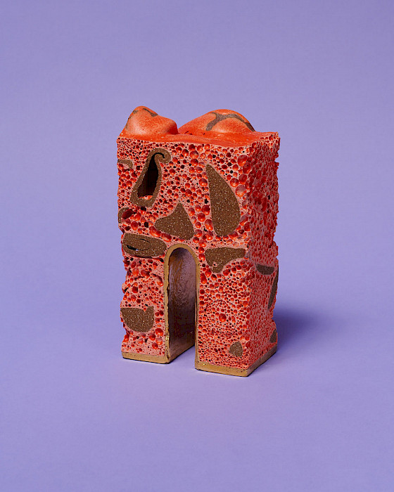 Photo of a red ceramic sculpture with brown spots. The u-shaped bottom of the sculpture forms a tall arch finished with gold.