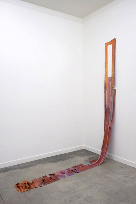 A thin, long piece of red-orange canvas hanging on a white wall cut and torn with half the canvas laying on the concrete floor.