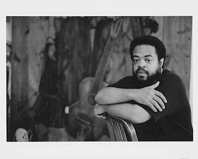 Black and white photo of a Black man sitting on a chair in front a guitar and coats hanging on a wood paneled wall.