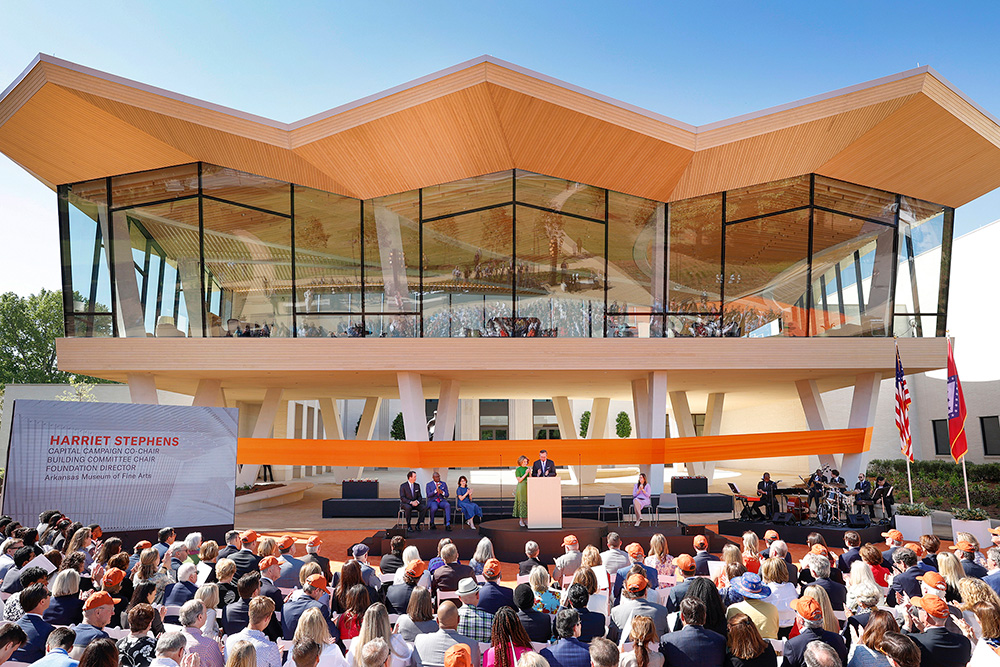 Photo of the ribbon cutting ceremony at the Arkansas Museum of Fine Arts. A large crowd sits facing a stage where two people are speaking at a podium. Behind them is a large orange banner extending from one side of the building to the other.
