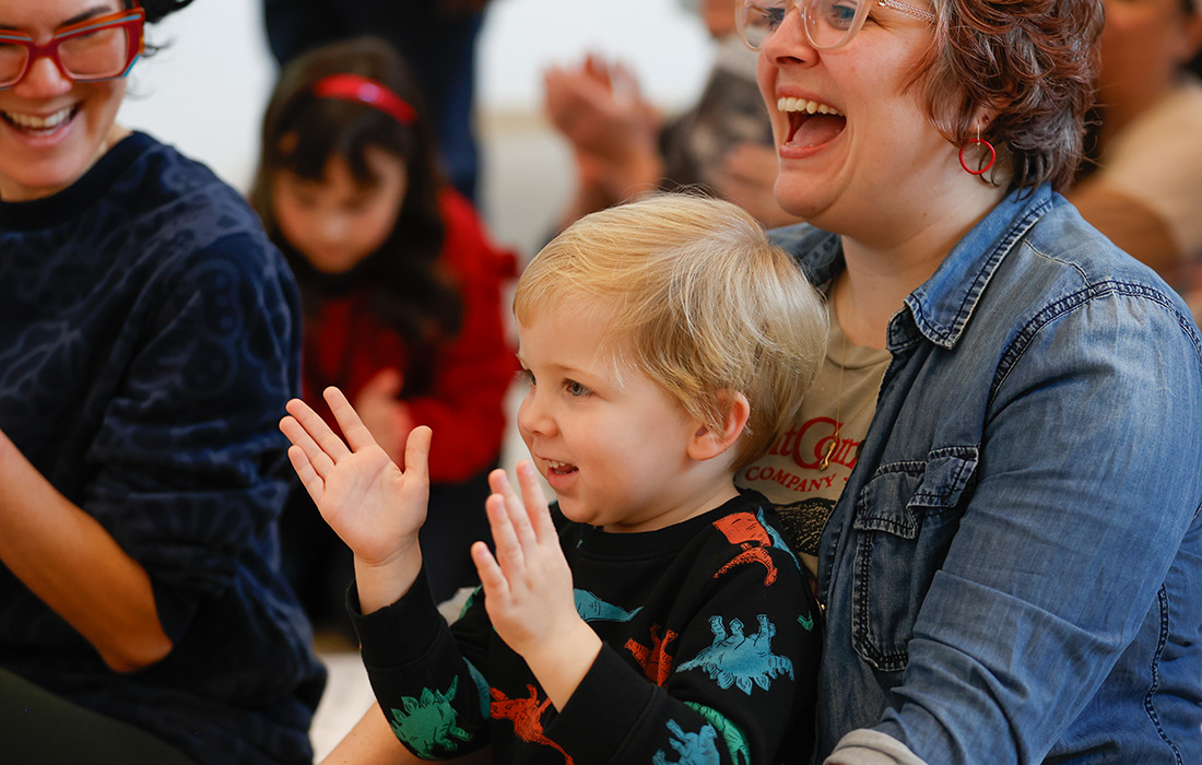 Photo of a toddler seated in his mother's lap and clapping while surrounded by other people.