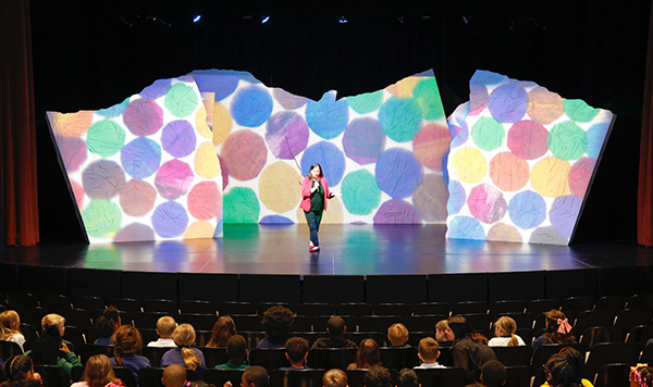 Photo of a woman standing on a theater stage in front of children in seats with a colorful stage background behind her.