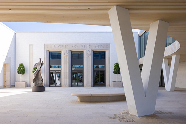 Photo of the Courtyard Entrance to AMFA viewed from underneath the Cultural Living Room. There is a semicircle concrete bench on the right and a large bronze abstract figure scuplture on the left.