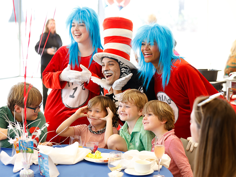 Photo of children seated at a table smiling and posing with actors dressed as the Cat in the Hat, Thing 1, and Thing 2 standing behind them.