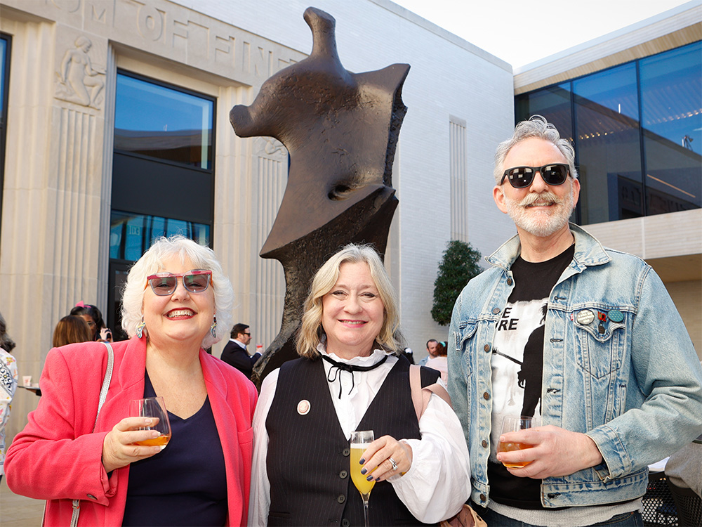 Photo of two middle aged women and a middle aged man holding cocktails and smiling while posed in front of a bronze sculpture in an outdoor courtyard.
