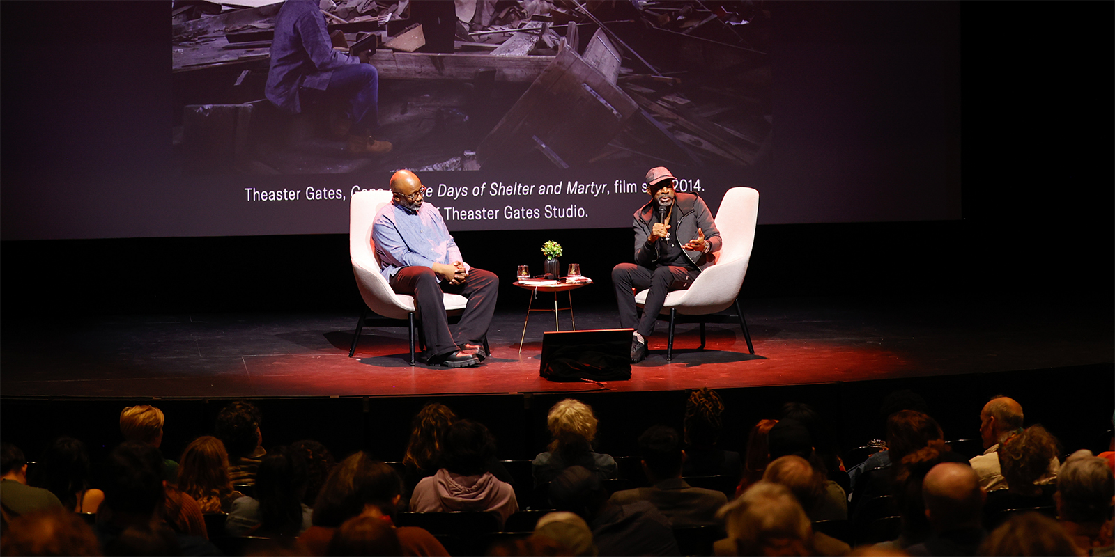 Photo of two men sitting in chairs on a dark stage. The man on the right is speaking into a microphone and gesturing towards the audience.