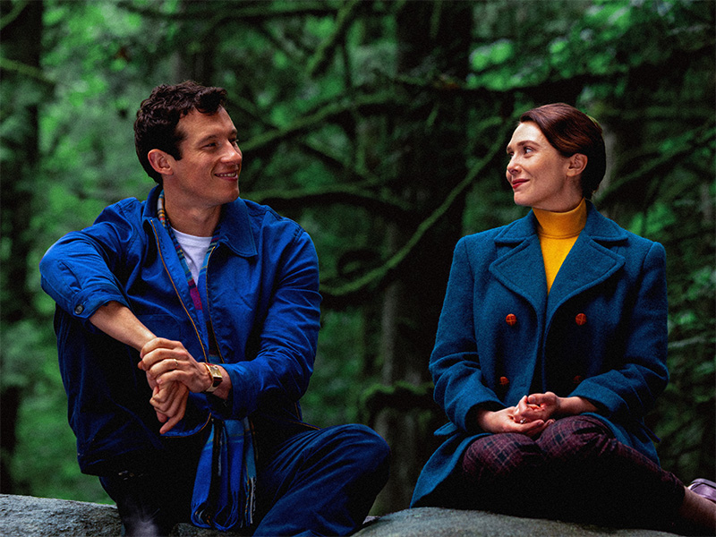 Film still depicting a man and a woman wearing jackets and sitting on a large rock in a forest.