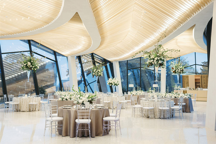 Photo of a room with floor to ceiling windows and an angular wood paneled ceiling. The room is decorated with a series of round tables, flowers, silvery tablecloths, and white chairs.
