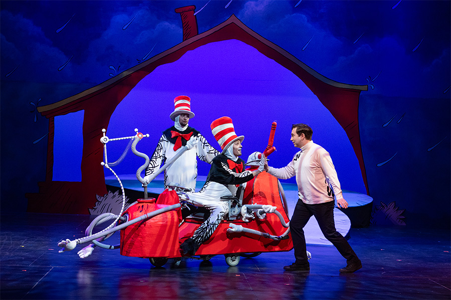 Photo of three people performing on a stage. Two of them are dressed as cats from The Cat in the Hat and a driving a cartoonish car.