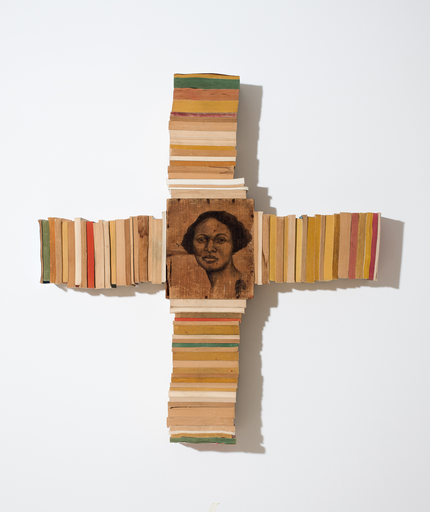 Photo of a drawing of a woman on a piece of wood set in the center of a horizontal line and vertical line of paperback books.