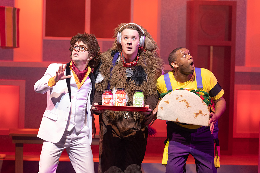 Photo of three men performing on stage. One is dressed in a 1970s period costume. One is dressed as a dog and holding a tray with three bottles of hot sauce. The other is dressed as a young boy and holding a giant taco.