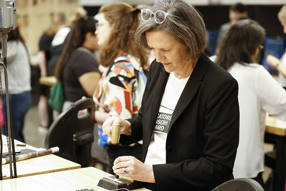 Photo of a woman with a short gray bob wearing a white t-shirt and black blazer. She is hammering metal in an art studio with other people in the background.