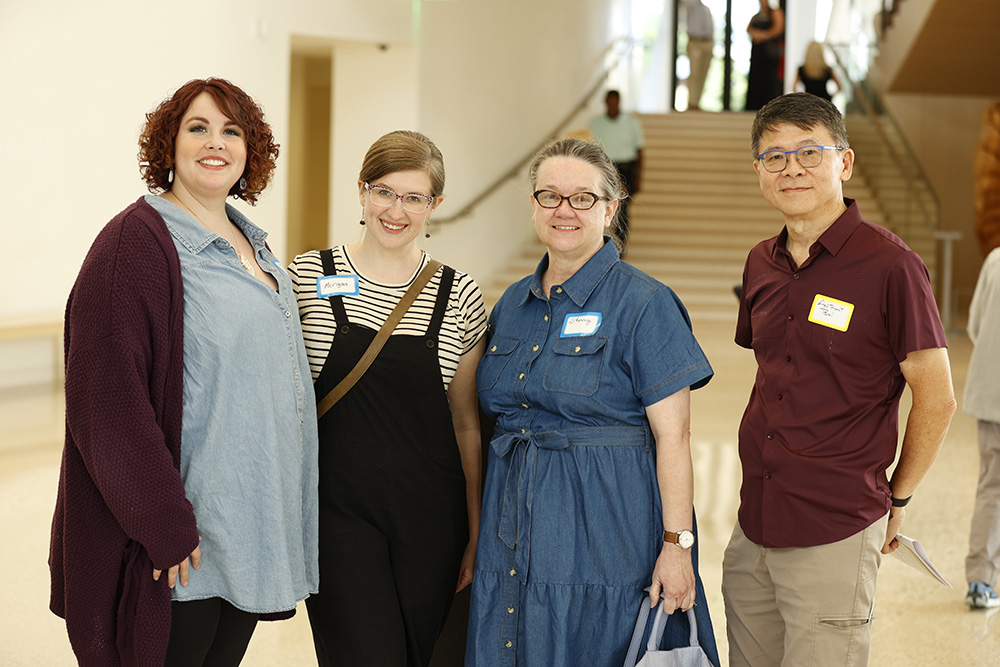 Photo of a three women and one man posed smiling together in the Atrium at AMFA. All four people are smiling and wearing name tags.